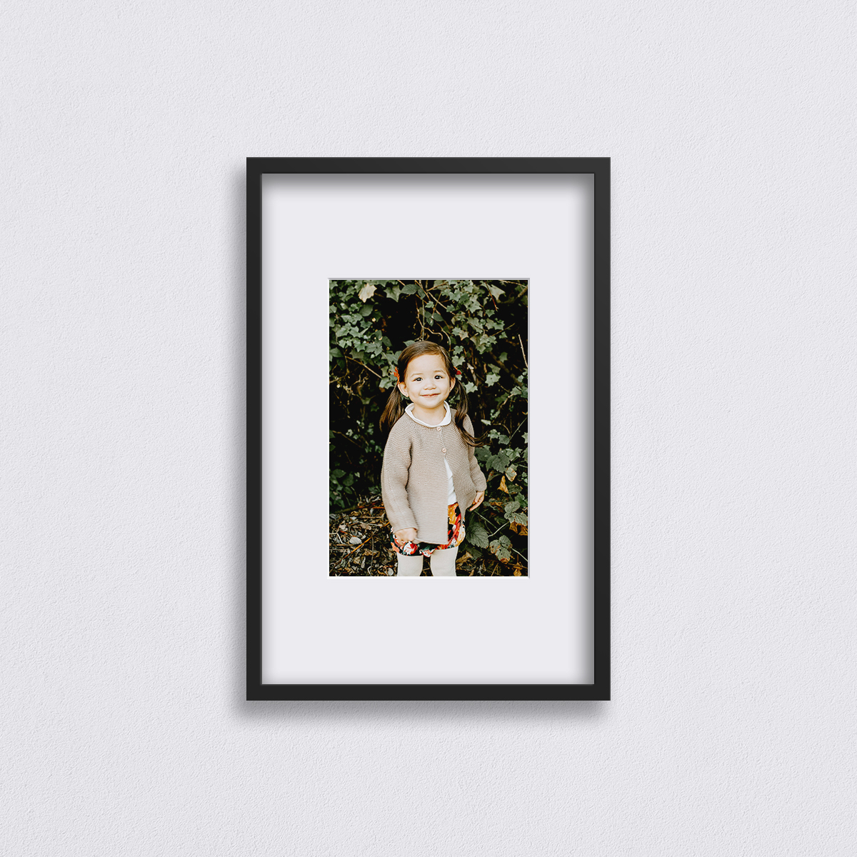 20 x 30 inch Artifact Uprising Deep Set Frame in Black finish matted to 12 x 18 inch featuring portrait of pig-tailed little girl in cardigan standing in front of background of green foliage