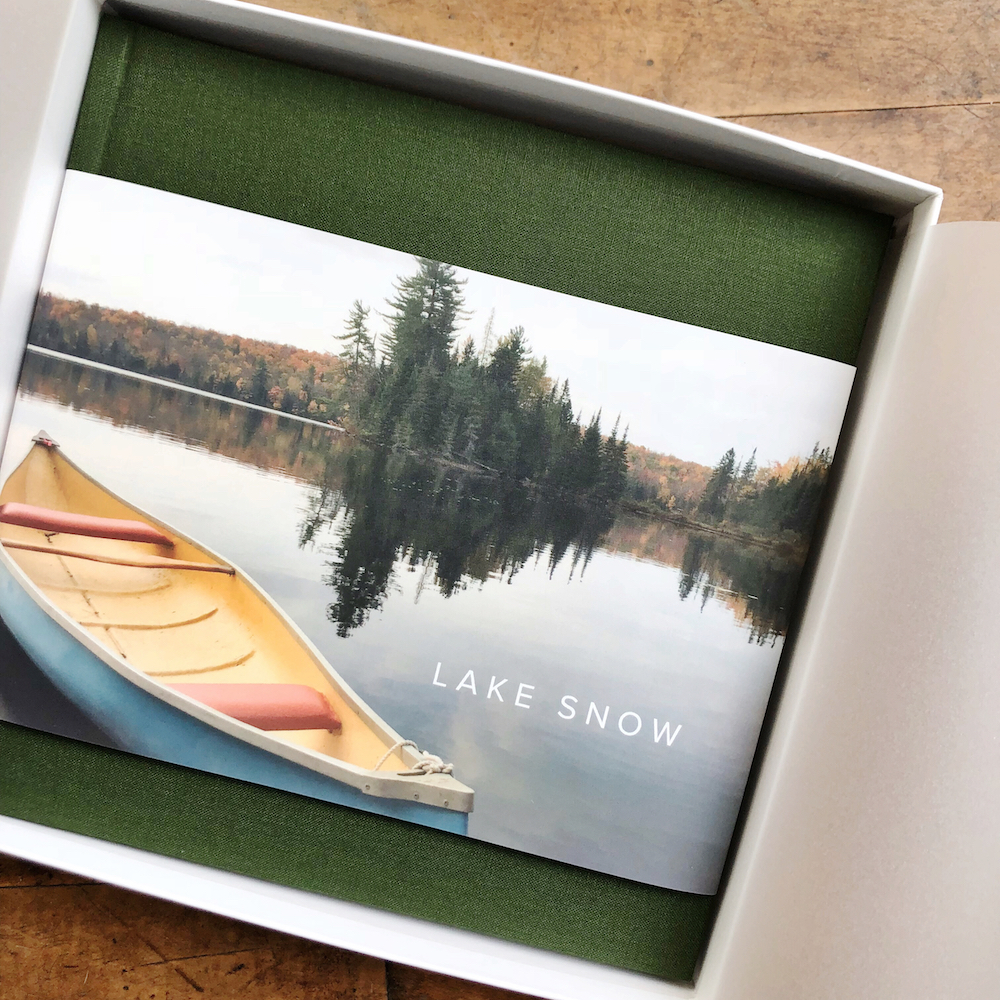Open white box revealing Artifact Uprising Hardcover Photo Book featuring photo of canoe at scenic lake