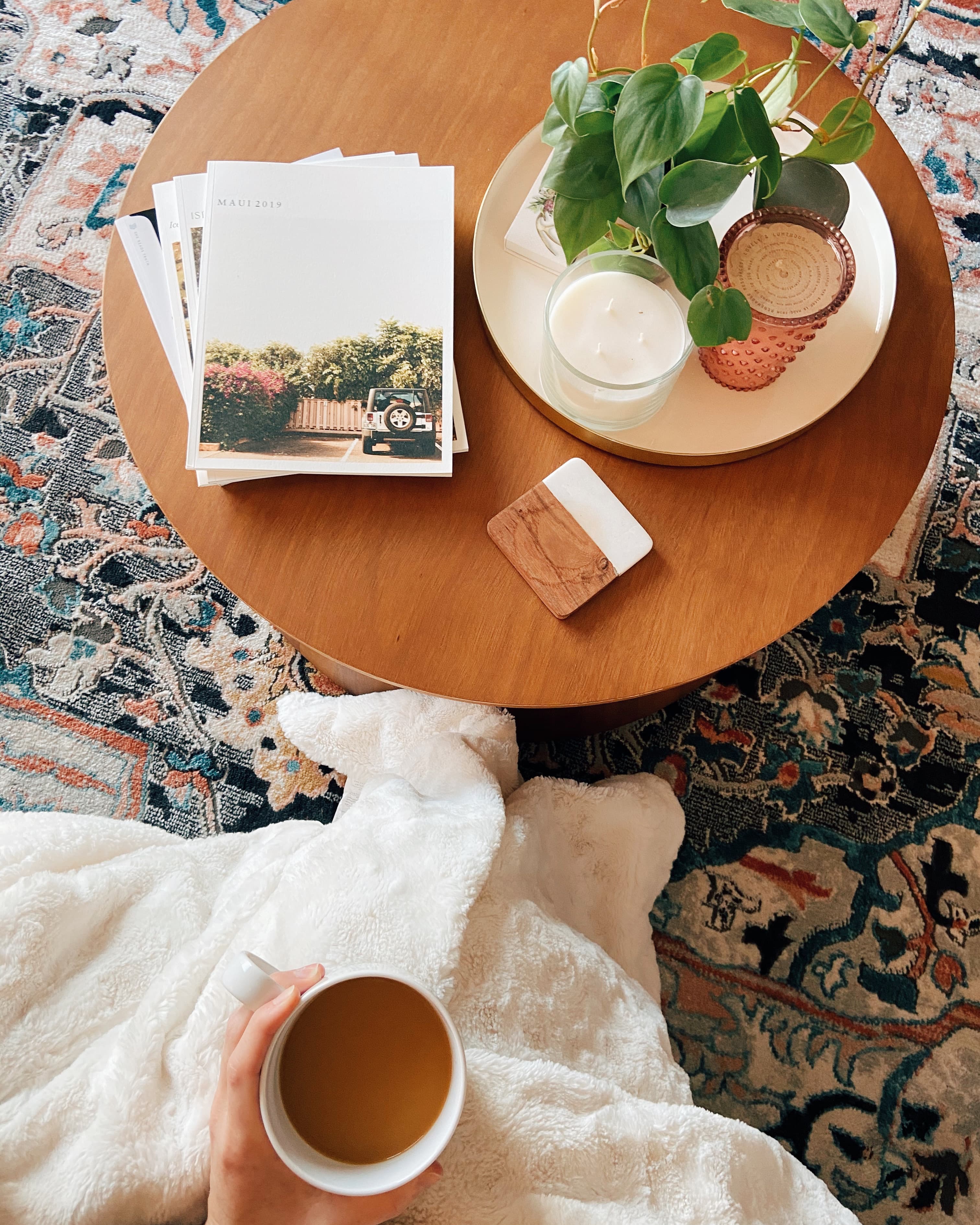 Styled photo of books on a table alongside a plant with a vibrant rug in the background. Female holding cup of coffee alongside fluffy blanket.